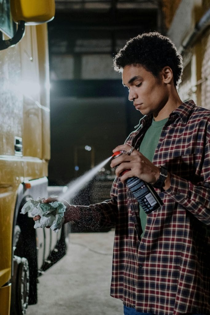 Young man using spray can to clean a vehicle in a garage setting.